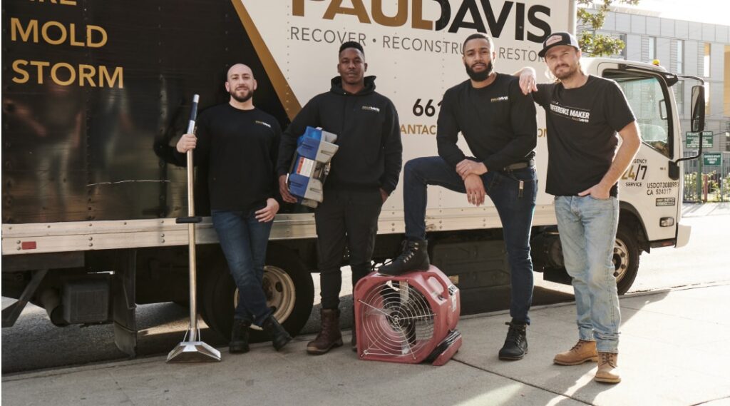 Paul Davis Restoration team members standing in front of a branded service truck with restoration equipment, including a floor squeegee, air mover, and drying tools, ready for water, mold, and storm damage response in Greater Missoula.
